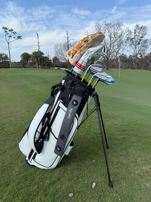 A white golf bag with black accents stands on grass, holding clubs with colorful headcovers and a Gray Magnetic Golf Cart Strap Sleeve featuring neoprene rubber, dual accessory magnets, velcro glove holder, and carabiner attachment. Trees and a flagstick are in the background.