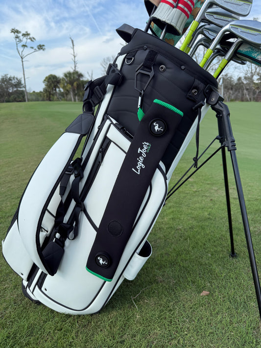 A Black/Green Magnetic Golf Cart Strap Sleeve made of neoprene rubber with dual accessory magnets, glove holder, and carabiner bag attachment is shown on a golf bag standing on green grass at a course with trees in the background.