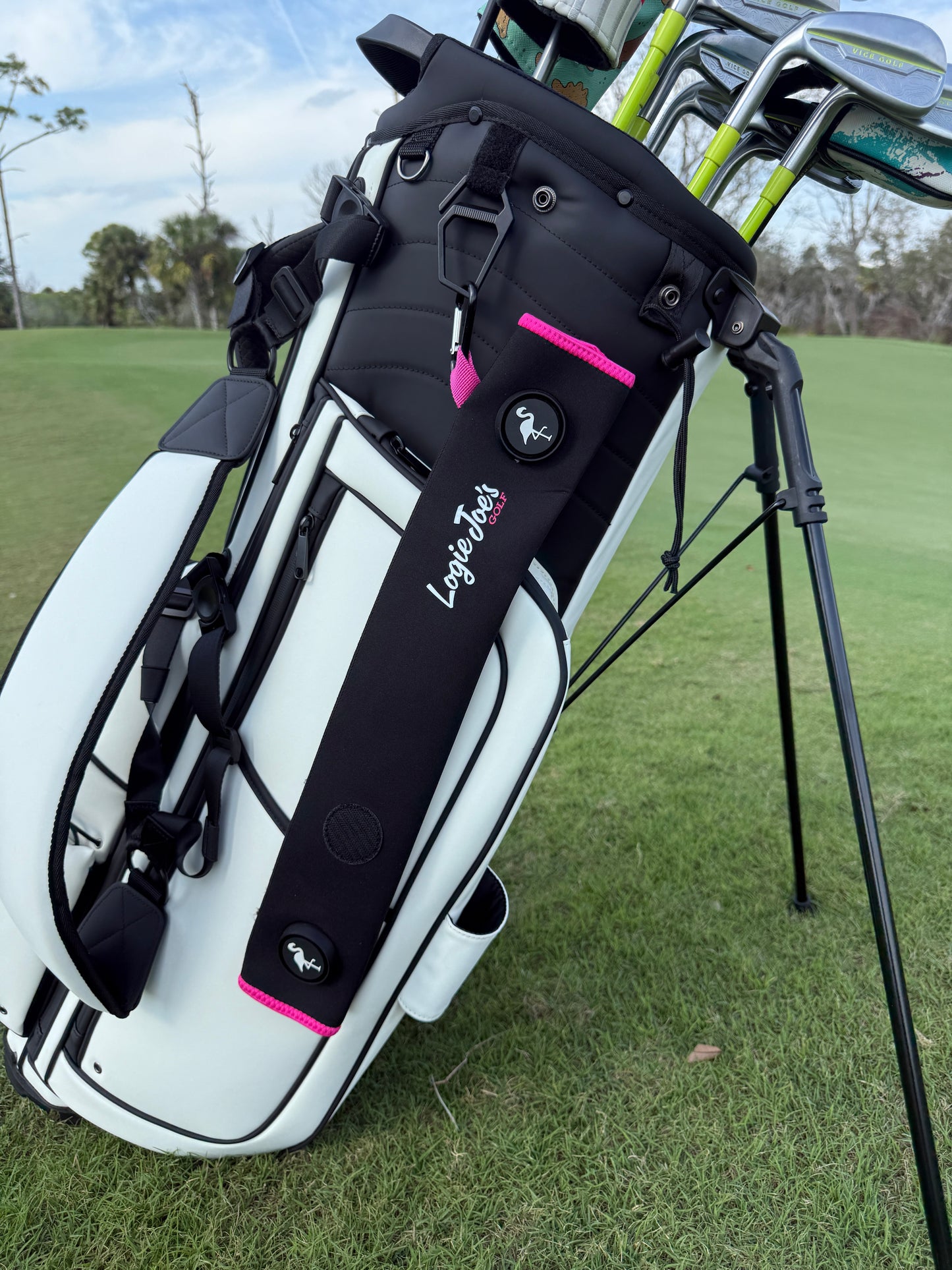 On green grass, a white and black golf bag holds clubs. Attached is a Black/Pink Magnetic Golf Cart Strap Sleeve (18.5 x 3.5 in, neoprene, dual accessory magnets, glove holder, carabiner). Trees and sky are visible in the background.