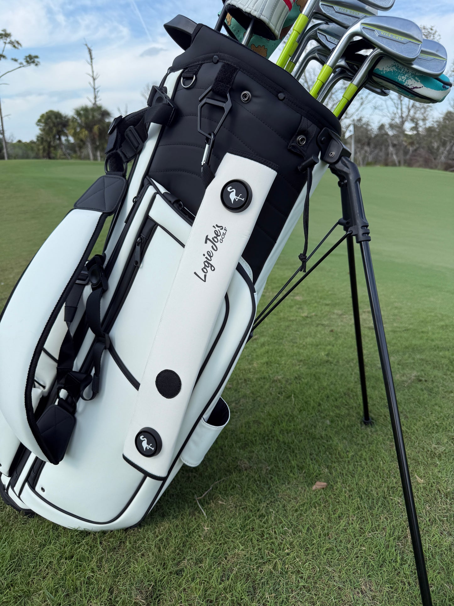 A White Magnetic Golf Cart Strap Sleeve by Loyola Tools, made of neoprene rubber with dual accessory magnets, a glove holder, and a carabiner bag attachment, is displayed on the grass near a golf green and trees.