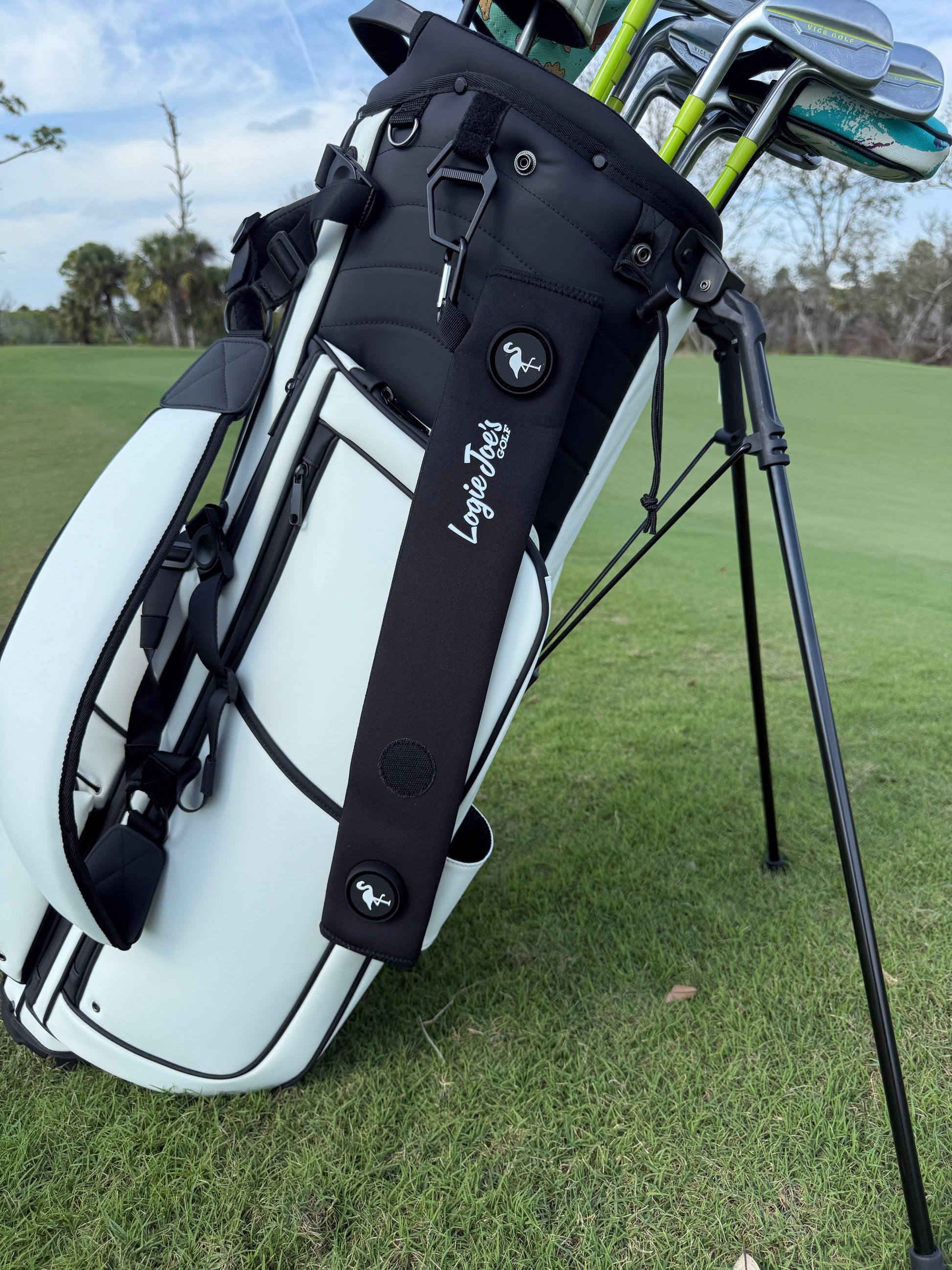 A white golf bag with clubs stands on green grass near a course. The Black Magnetic Golf Cart Strap Sleeve (18.5 x 3.5 in) features neoprene rubber, dual accessory magnets, glove holder, carabiner attachment, and small animal logos. Trees and blue sky are visible.