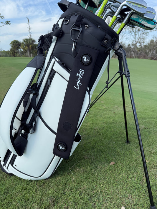 A white golf bag with clubs stands on green grass near a course. The Black Magnetic Golf Cart Strap Sleeve (18.5 x 3.5 in) features neoprene rubber, dual accessory magnets, glove holder, carabiner attachment, and small animal logos. Trees and blue sky are visible.