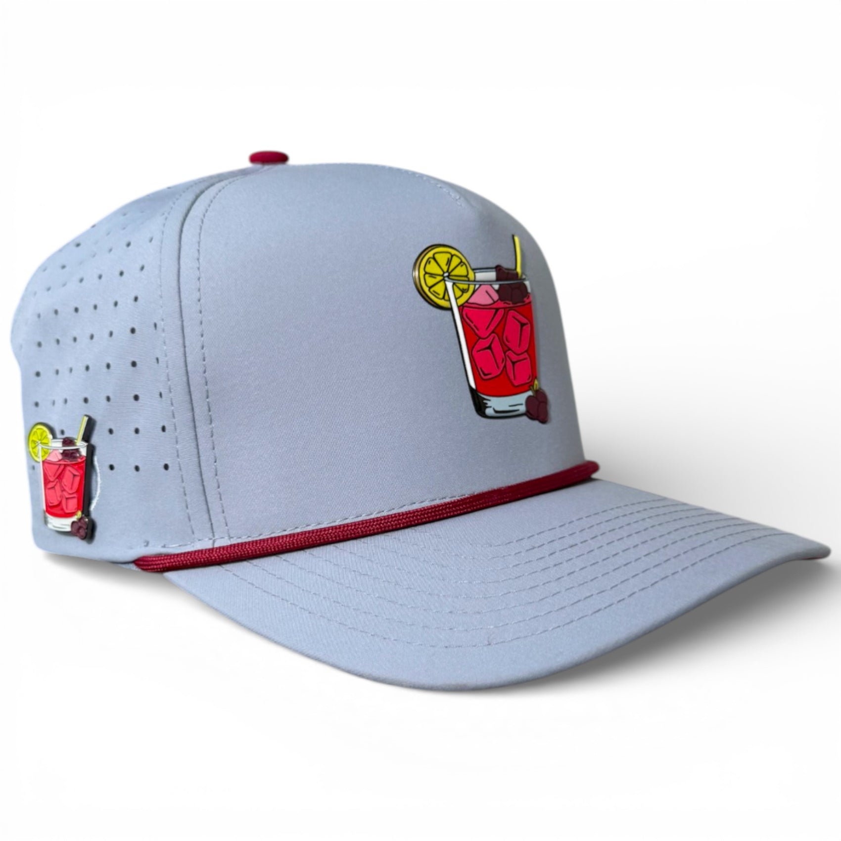 The Transfusion Golf Hat is a light gray snapback cap with an illustrated patch of a red iced drink and lemon slice, maroon trim, side ventilation, adjustable fit, and includes a matching pin plus a built-in magnetic ball marker with holder.