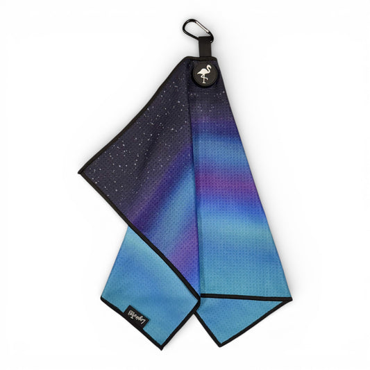 The Northern Lights Golf Towel features a blue and purple aurora gradient with a starry night design, black edges, a round flamingo logo patch, carabiner clip, and is crafted from double-sided microfiber. Shown on a white background.