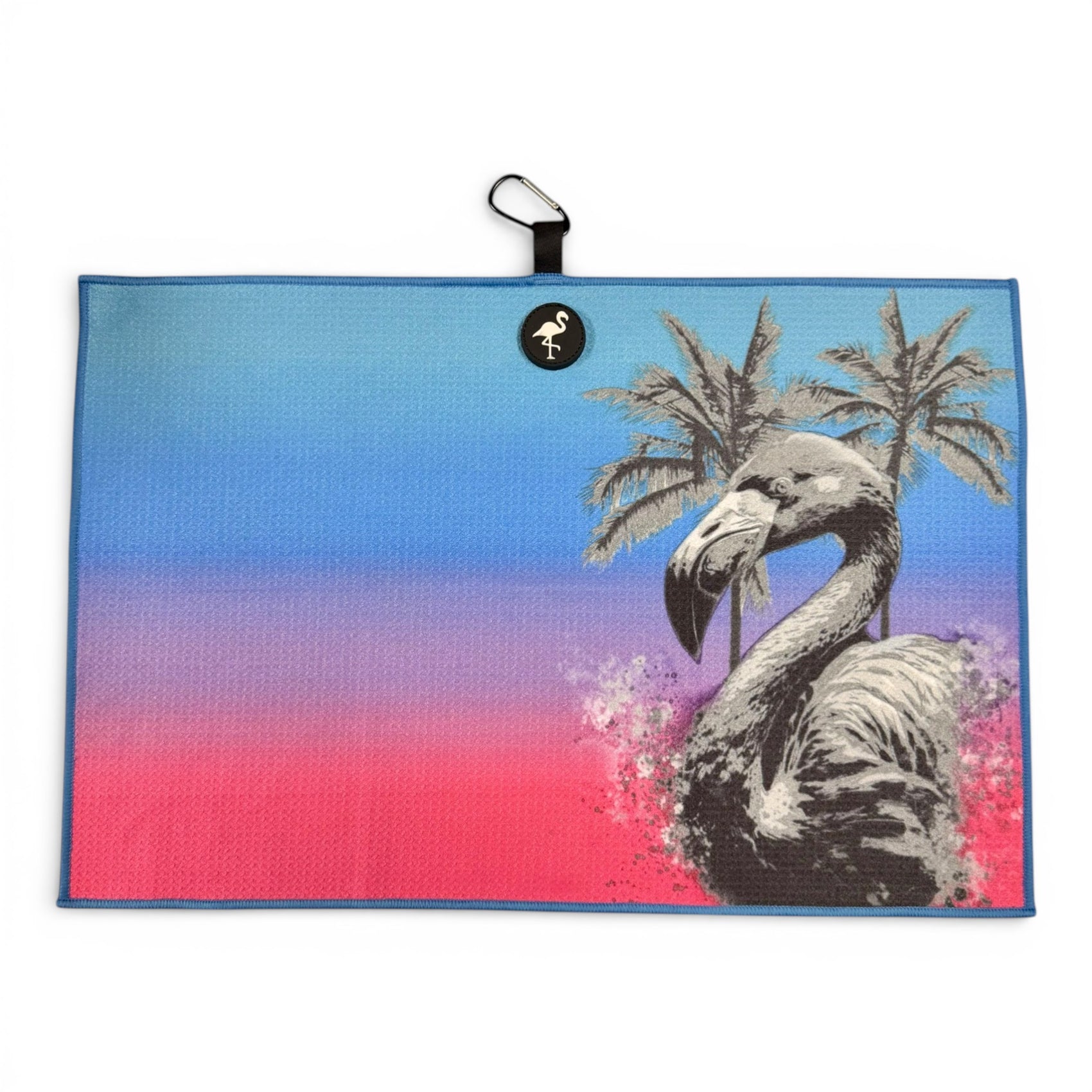 The Flamingo Palm Golf Towel features a vibrant blue-to-pink gradient, black and white flamingo and palm trees, a small flamingo logo, and includes a carabiner clip. Its premium double-sided microfiber is both fun and functional for golf games.