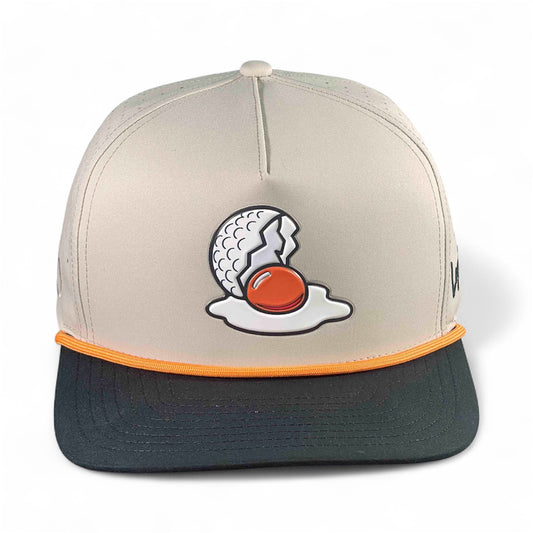 The Breakfast Ball Golf Hat in Mulligan Tan is an adjustable snapback cap with a built-in magnetic ball marker holder and marker. It features a beige and black design with golf egg embroidery—perfect for your next mulligan.